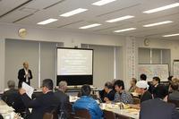 12月4日に行われた小規模多機能自治勉強会にて多くの人が席に座っている中で一人の人がスクリーンの隣で話している写真
