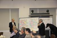 氷見市地域自慢大会の第1部にて数人が席に座っているのが写っている中で付箋が貼られたホワイトボードを用いて二人の人が話している写真