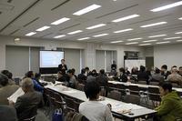 氷見市地域自慢大会にて室内で多くの人が席に座っている中で一人の人がスクリーンの前で立って話している写真