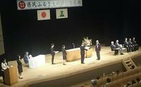 県民会館で開催された記念式典のステージで賞状を手渡される様子の写真