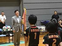 市長がマイクの前に立ち向かい合ってバレーボールのユニフォーム姿の児童達がならんでいる体育館の写真