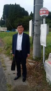 田んぼ脇の道路にあるバス停の横に立つ市長の写真