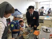 エプロンを着た男の子が、菜箸を鍋に入れて料理をしている様子を市長が見守っている写真