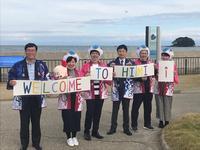 富山湾を背に「WELCOME TO HIMI!」と書かれたボードを持つ市長達の写真