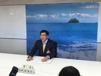 島が見える海の写真の前に着席し記者会見をする市長の写真