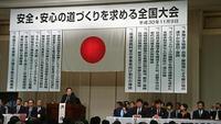 文字が書かれた垂れ幕と日本国旗の印が飾られたステージで複数の男性が座り、中央の男性が立っている写真
