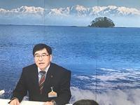 定例記者会見にて青空の広がる海に浮かぶ小島がプリントされた垂れ幕を背に答弁をする市長の写真