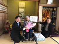 菅田氏のご自宅の床の間の前でお祝いを手にした市長と並んだ菅田氏と男性がいる写真