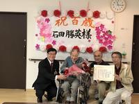 「祝百歳」と書かれた壁掛けボードの前で石田氏に花束を手渡す市長の写真