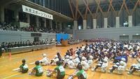体育館でスポーツ団体方々が床に座り総合開会式が行われている写真