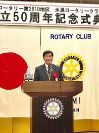 記念式典で日本の旗とロータリークラブの旗の前の演壇に立つ市長の写真