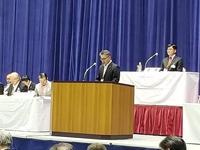 市長会総会のステージ上の演壇で発言する方が立つ演壇後ろの議長席に着席した市長の写真
