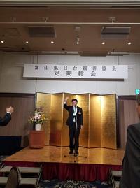 定期総会のステージ上の金屏風の前に立ち右手をあげる市長の写真