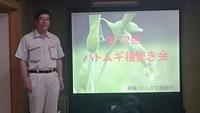 第12回ハトムギ種まき会と投影されたスクリーンの左に作業服姿で立つ市長の写真