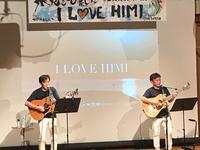 「ILOVE HIMI」と書かれている看板の前でアコースティックボーイズがギターを弾きながら歌っている写真