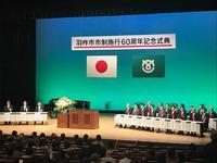 日章旗と市のマークの旗が掲示されたステージで左右のテーブルに関係者や来賓の方々が着席している記念式典の写真
