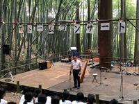 竹林の中で竹ドームコンサートと書かれたステージに立つ男性の写真