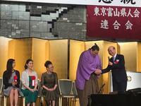 東京富山県人会連合会の懇親の集いで3人の芸能人が着席し立って朝乃山関が握手している写真
