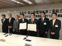 関係者7名が一列に起立し市長と富山県電気工事工業組合の方が協定書を見せている写真