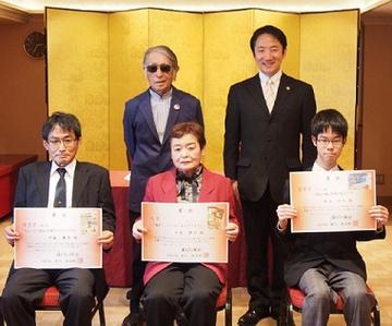 二人の男性と一人の女性が表彰式で賞状を持って藤子(A)先生や本川市長と記念撮影している写真